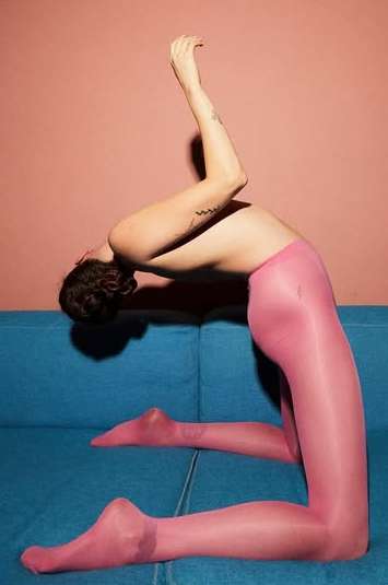 Adult kneeling on blue couch wearing pink tights with arms raised in a studio with peach backdrop; head bent backward.