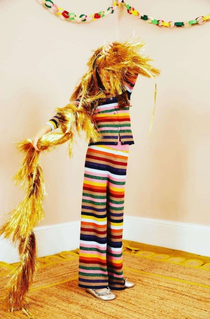 A child in striped pants and top stands in a corner, obscured by tinsel, under a chain garland. Bright, playful fashion photography.