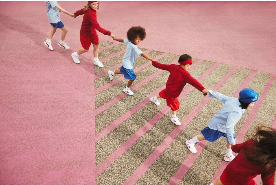 Kids in red and blue athletic wear holding hands and walking on a light pink and tan patterned surface in an overhead shot.