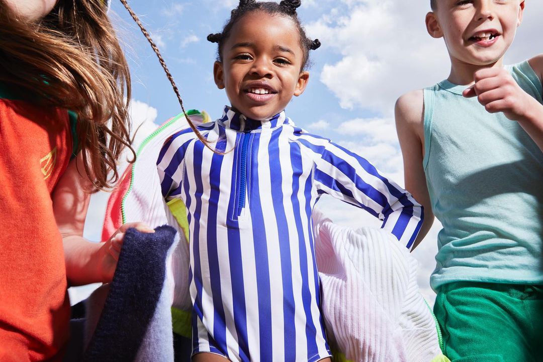 Three children are standing outdoors on a sunny day, one is wearing a blue and white striped swimsuit, smiling at the camera.