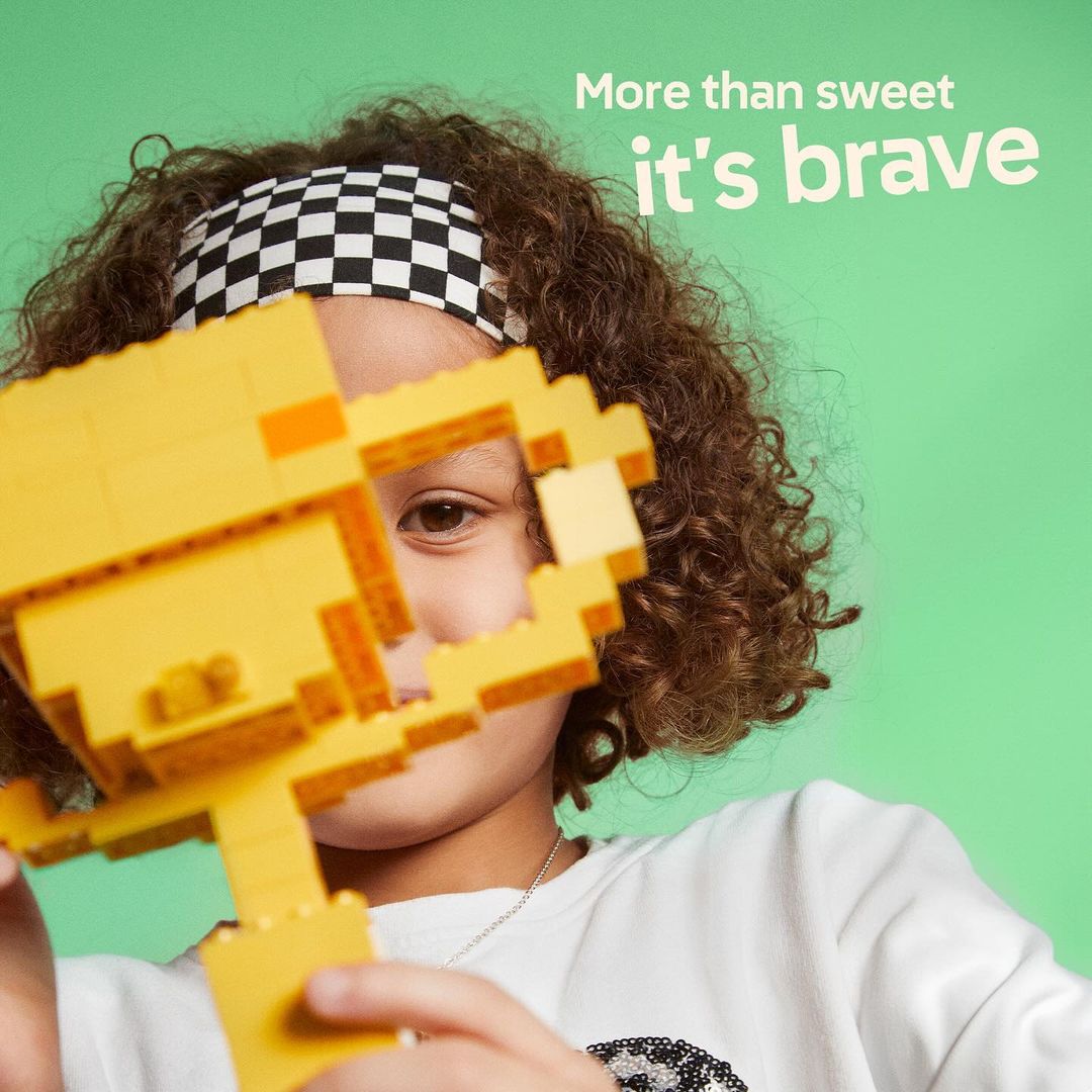 Young child holding a yellow toy block trophy with checkerboard headband, against a vibrant green background. Studio shot, eye-level.