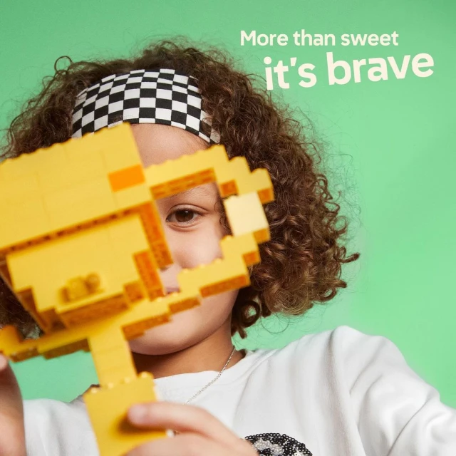 Young child holding a yellow toy block trophy with checkerboard headband, against a vibrant green background. Studio shot, eye-level.