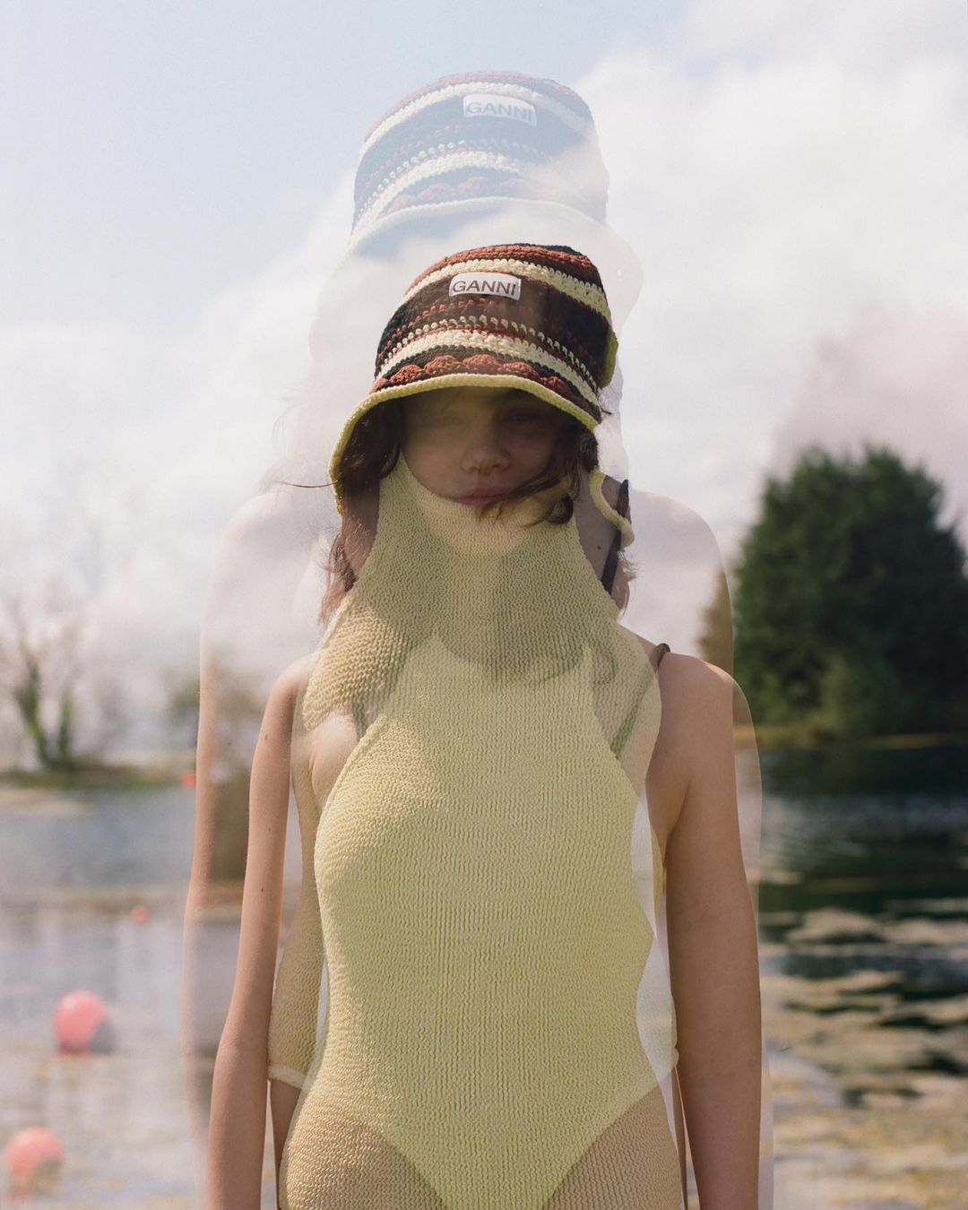 A woman wears GANNI knitted bucket hat and swimsuit outdoors, with a blurred, layered effect creating a dreamy fashion image.