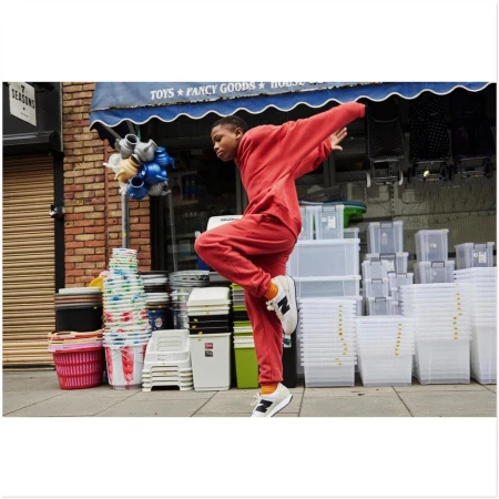 Young person in red sweatsuit strikes a dynamic pose on a city sidewalk with storefronts and stacked goods as backdrop, creating an energetic street style scene.