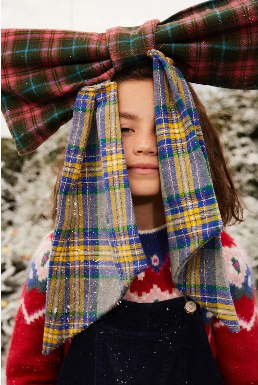 Young girl with plaid hair bow and sweater poses indoors against a snow-like background, creating a festive holiday fashion portrait.