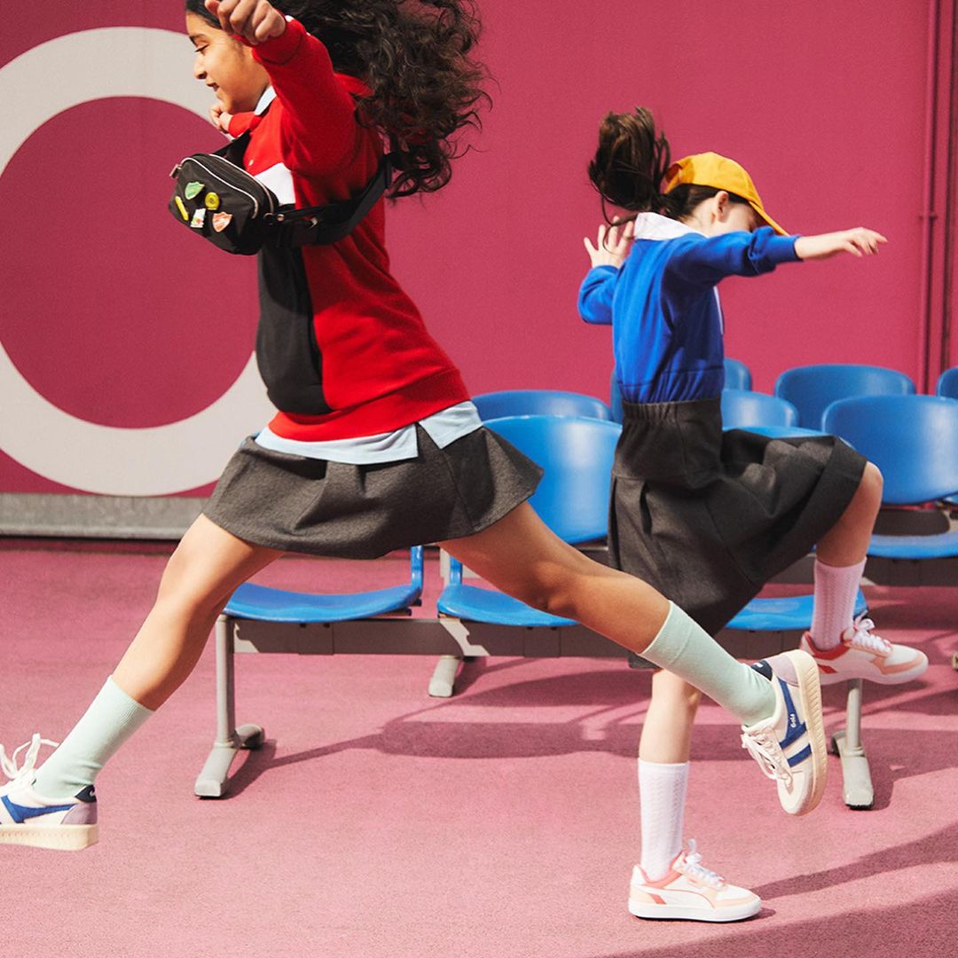 Two children joyfully jump in a pink room with blue chairs, one in red and black, the other in blue and yellow school uniforms.