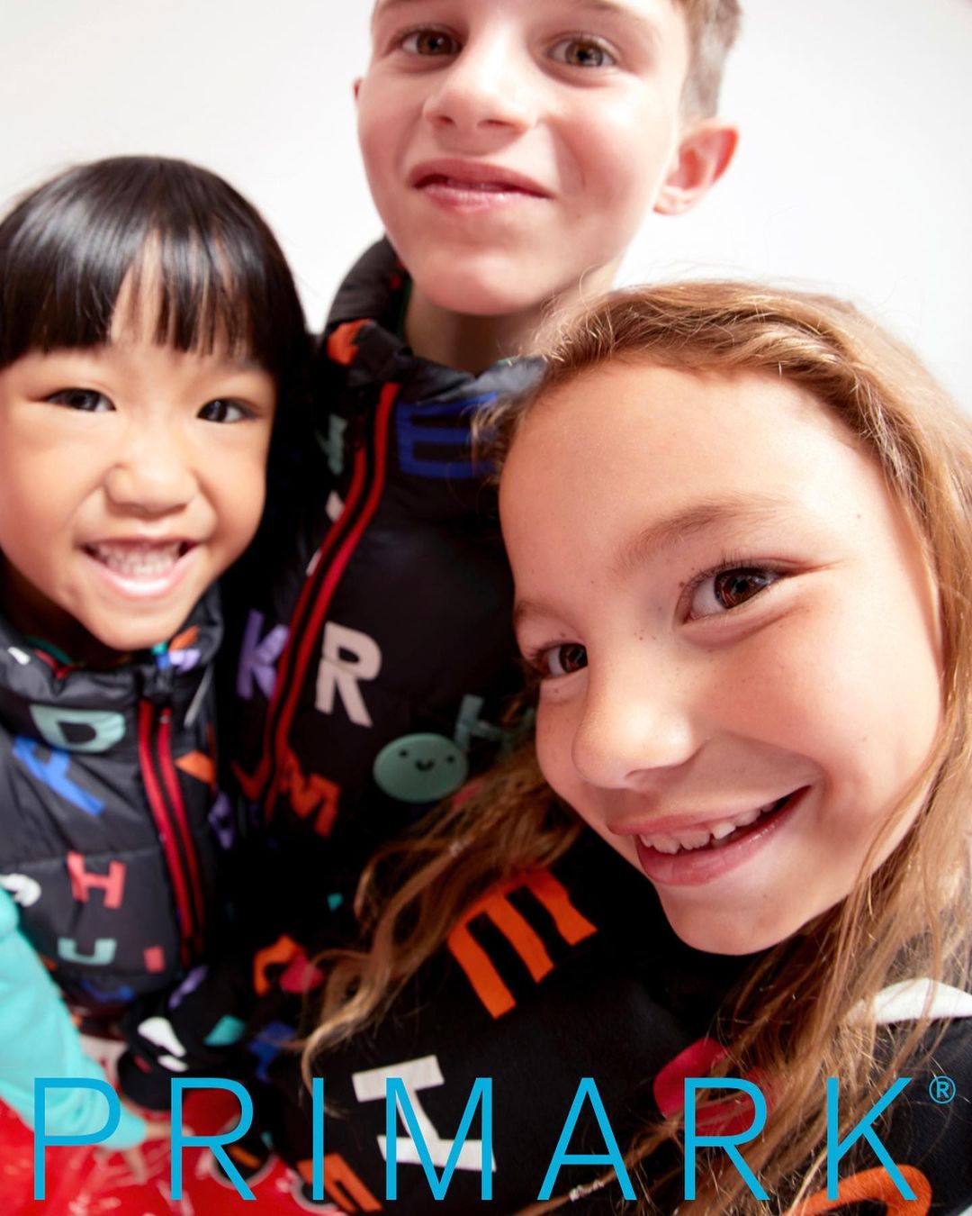 Three smiling children in letter-printed puffer jackets pose close together indoors. The clothing brand Primark is visible at the bottom.
