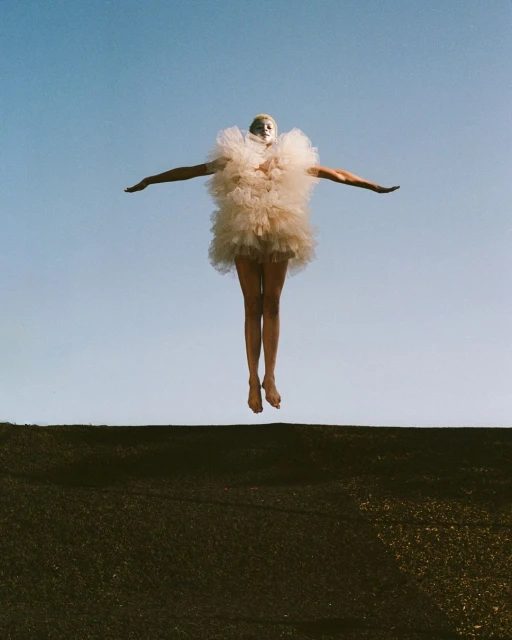 A person in a fluffy, ruffled dress jumps against a blue sky, arms outstretched, balanced above a dark wall, captured in a low-angle shot.
