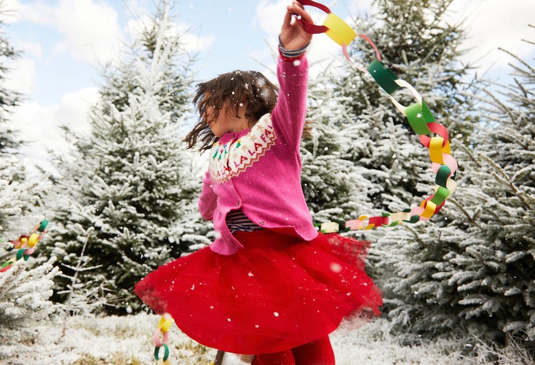 A young girl twirls in the snow, wearing a pink cardigan and red tulle skirt, amidst snow-covered fir trees and holding a colorful paper chain.