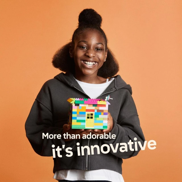 Happy African American girl smiling and holding up a colourful building-block model house on a solid orange background.
