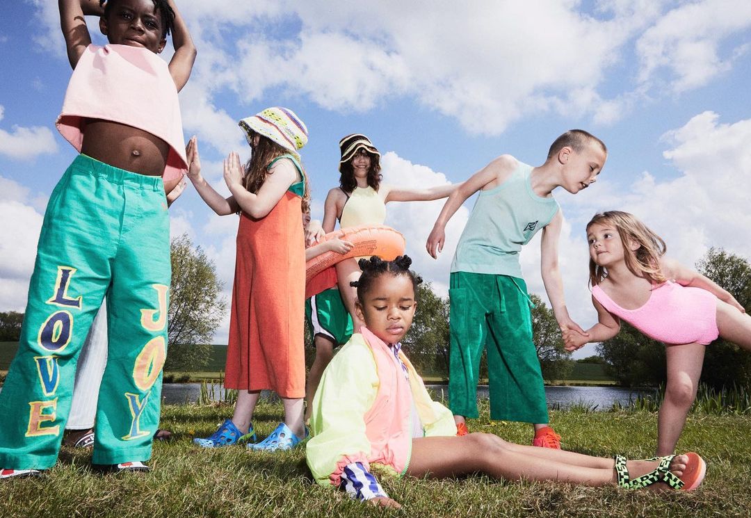 A group of seven children wearing colorful summer clothing are standing and sitting in a grassy field on a sunny day with a blue sky.