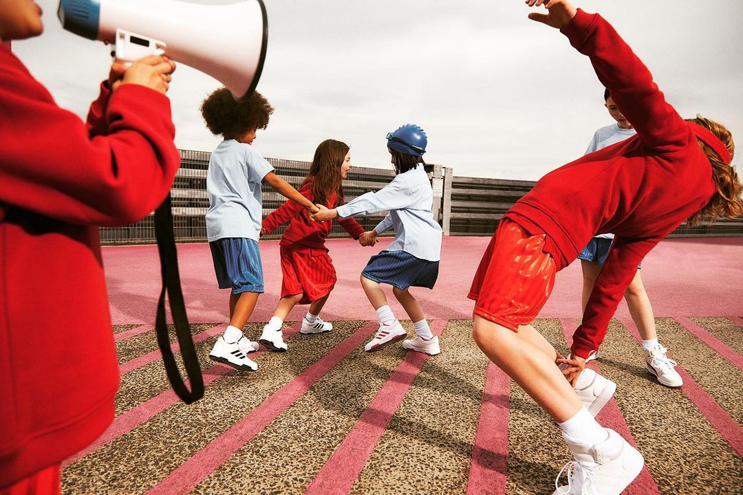 Children in red and blue athletic wear play Ring Around the Rosie on a striped pink surface with a person holding a megaphone nearby.
