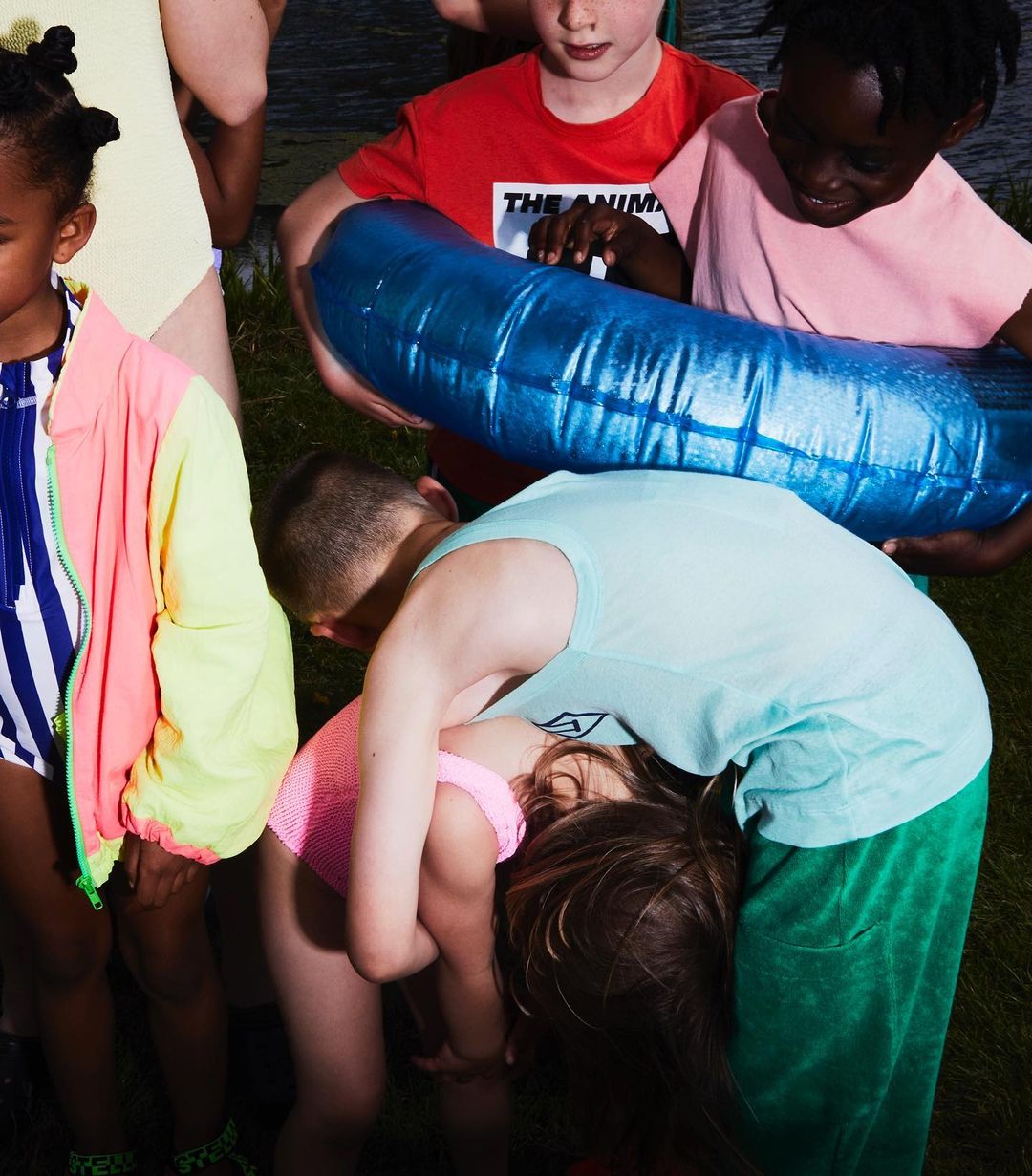 A group of children in swimsuits and casual clothing gather outdoors near water with an inflatable pool toy on green grass.