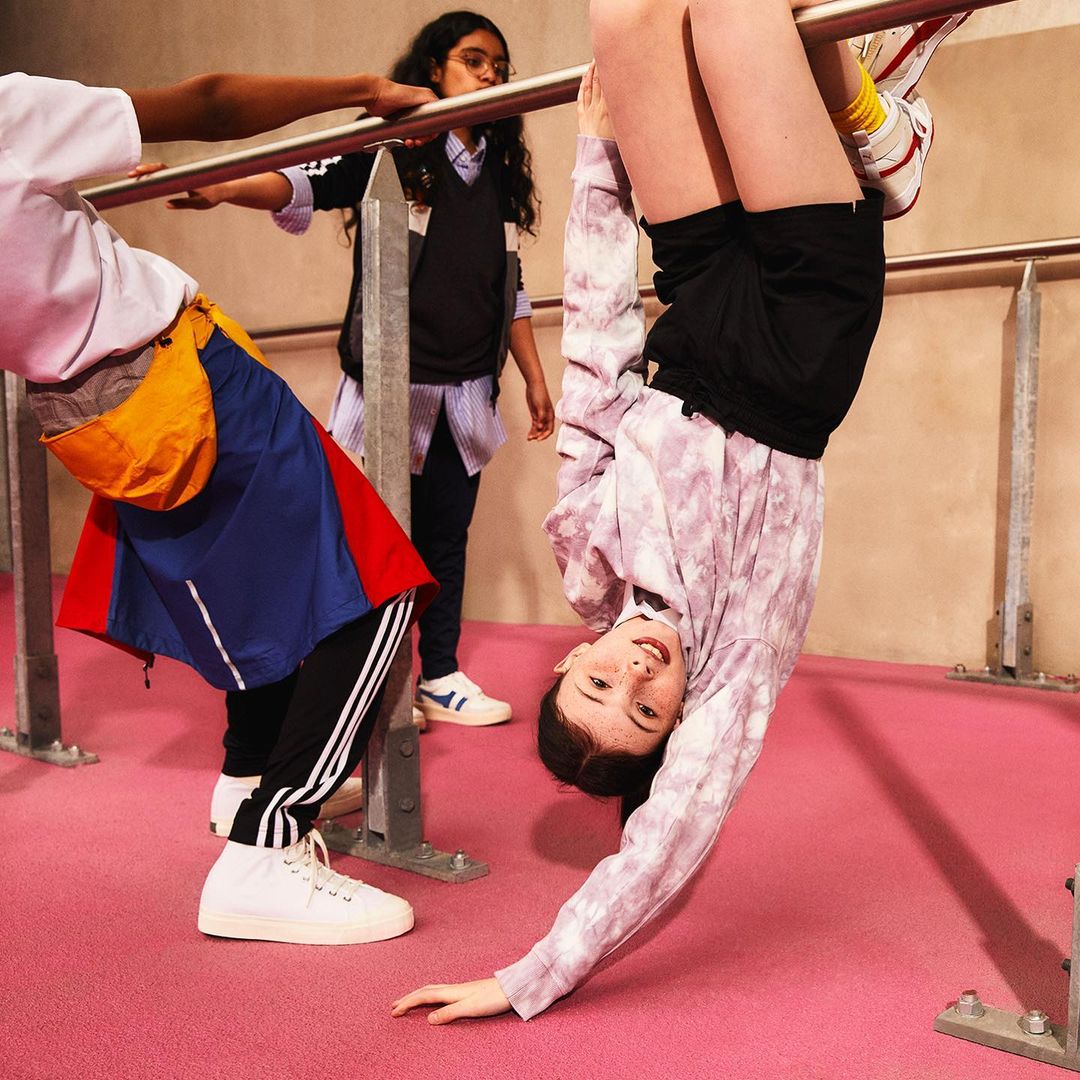 Upside-down view of a tween girl hanging from a bar with friends, wearing a patterned sweatshirt and shorts, in an indoor location.