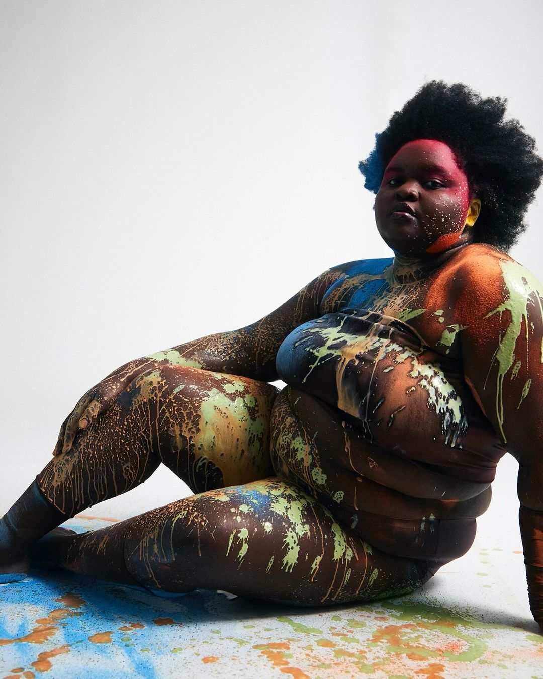 Portrait of a Black woman sitting with paint on her skin in a studio setting. The woman has short, dark hair and is staring at the camera.