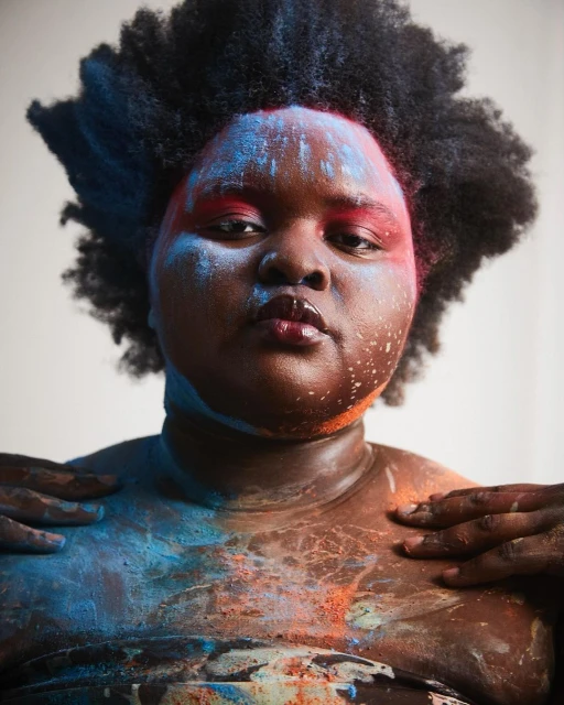 Portrait of a woman with dark skin, Afro hair, and blue and orange paint on her face and body against a simple light background.