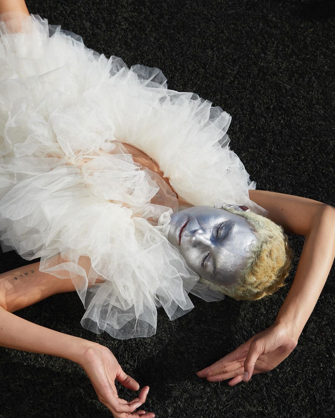 A woman with silver face paint lies on black surface, wearing a voluminous white tulle ruffled dress in an artistic fashion portrait.