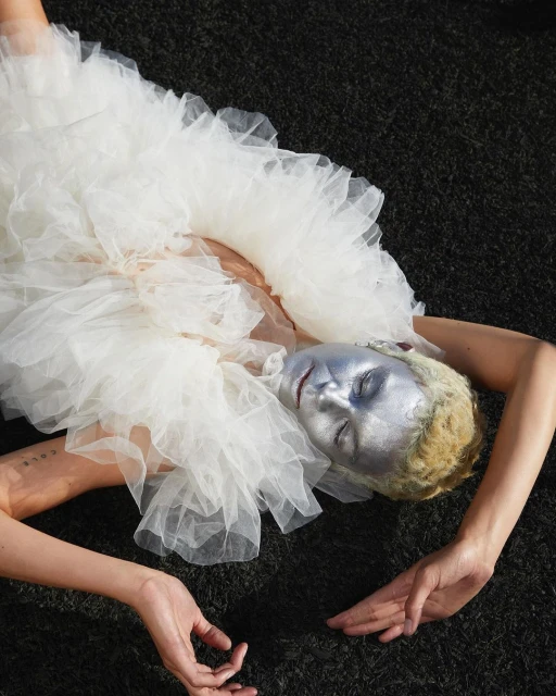 A woman with silver face paint lies on black surface, wearing a voluminous white tulle ruffled dress in an artistic fashion portrait.