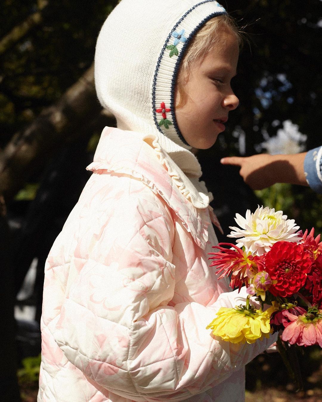 Young girl in a bonnet and quilted jacket holds a bouquet of colorful dahlias, standing outdoors in a natural setting with foliage.
