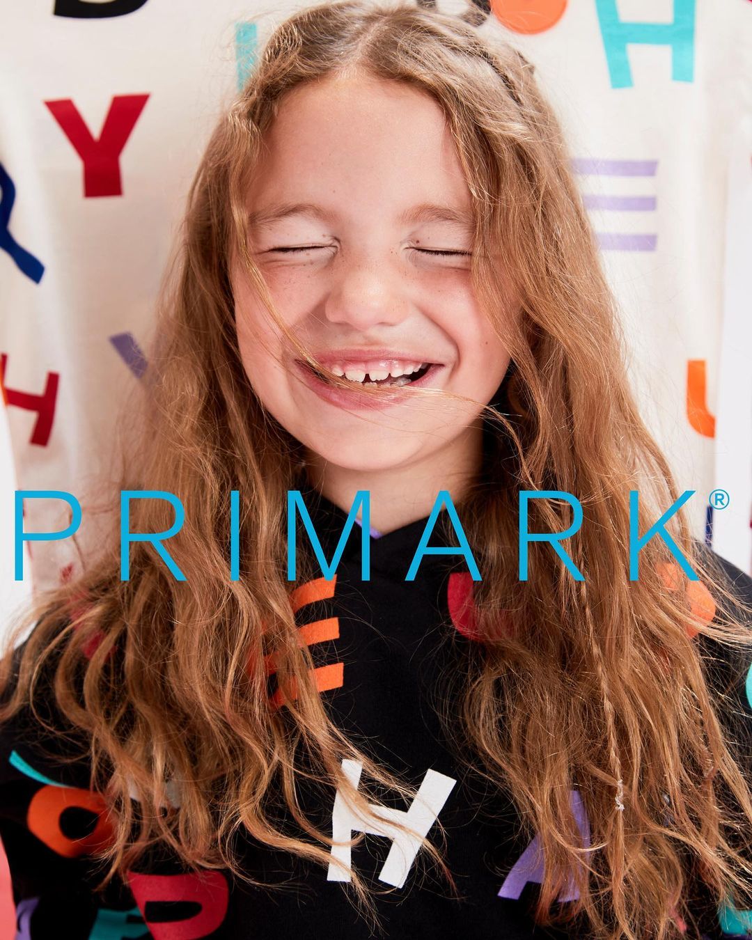 Close-up of a smiling young girl with long brown hair wearing a colorful printed sweatshirt; background features letters.