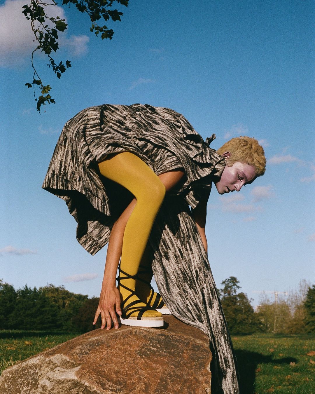 A person in a patterned dress and yellow tights poses on a rock in a grassy field under a blue sky, showcasing a fashion editorial.