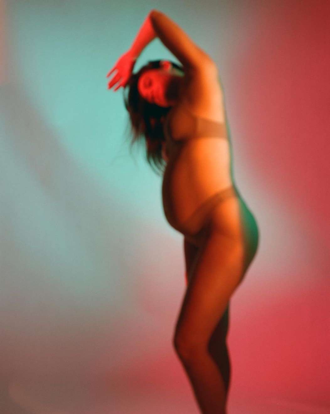Pregnant woman in underwear poses in a studio with red and blue lighting creating a blurred, dreamy effect emphasizing form and body.
