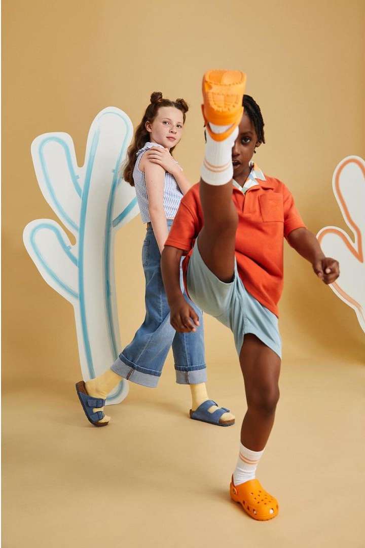 Two children pose in a photo studio with painted cactus props. One child has their leg up in the air showing off orange clogs.
