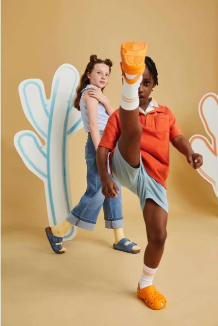 Two children pose in a photo studio with painted cactus props. One child has their leg up in the air showing off orange clogs.