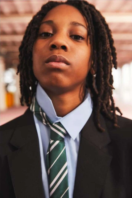 Portrait of a Black person with dreadlocks wearing a black blazer, light blue shirt, and a green striped tie, standing indoors.