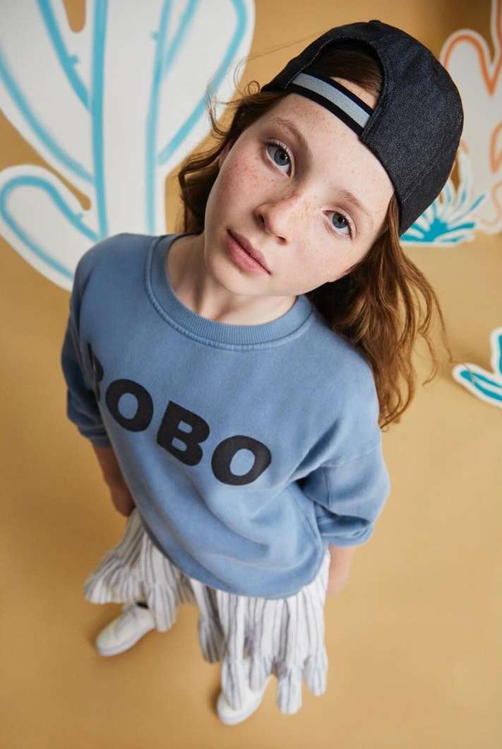Child wearing blue sweatshirt and dark baseball cap looking up in high-angle portrait by Nerys Jones