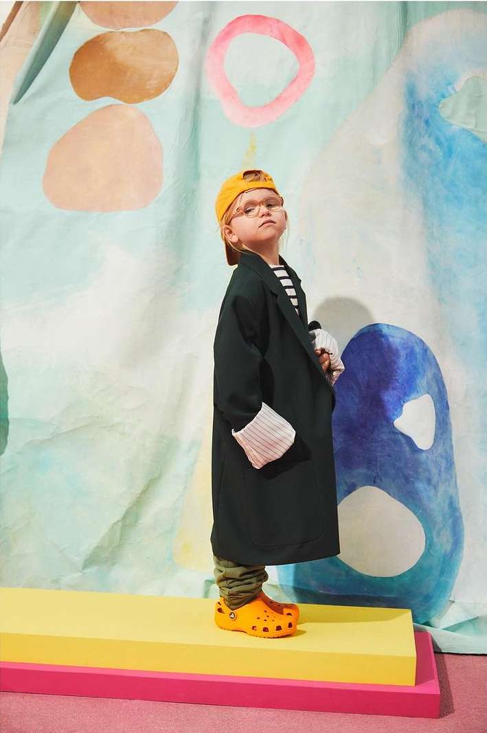 Child model wearing oversized blazer and yellow cap poses on colorful platform in front of a painted backdrop, showcasing fashion style.
