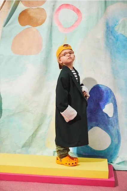 Child model wearing oversized blazer and yellow cap poses on colorful platform in front of a painted backdrop, showcasing fashion style.