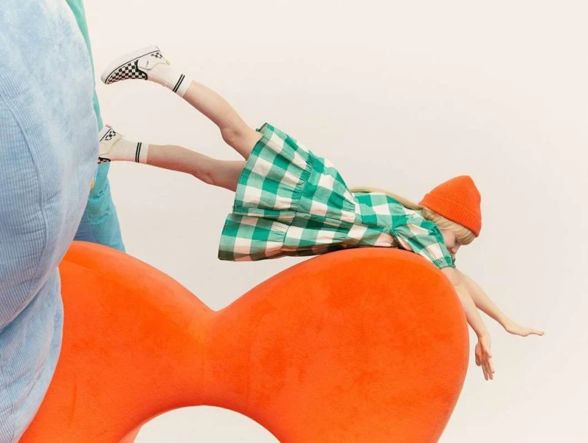 Child in green and white checkered dress and orange beanie hat lies on an orange prop in front of a white background in a studio setting.