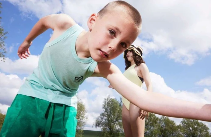 Young child in pale green tank top and green shorts reaching toward camera with another person in background