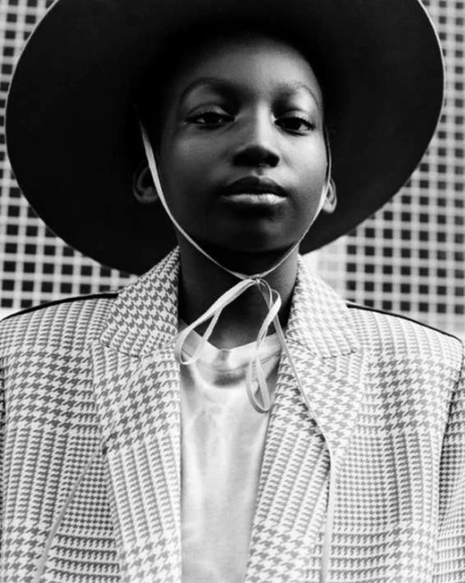 Monochrome portrait of a young Black person wearing a hat and patterned jacket, posing against a textured background, fashion forward.