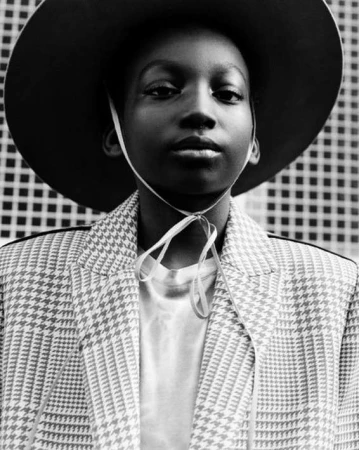 Monochrome portrait of a young Black person wearing a hat and patterned jacket, posing against a textured background, fashion forward.