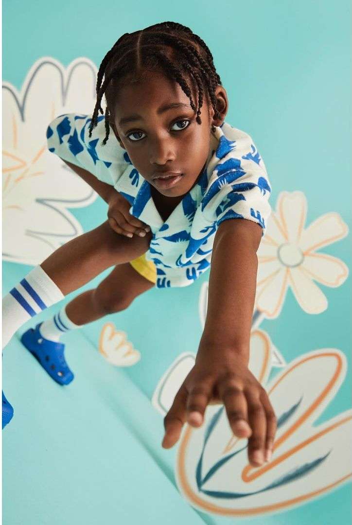 Young boy with braided hair, posing in a blue and white patterned shirt, yellow shorts, and blue clogs, against a floral backdrop.