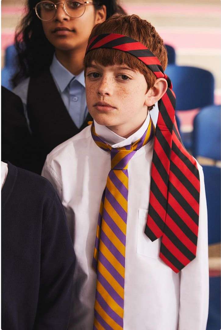 Young boy with red hair and freckles wearing a striped head band and a tie around his neck, standing in a classroom setting.