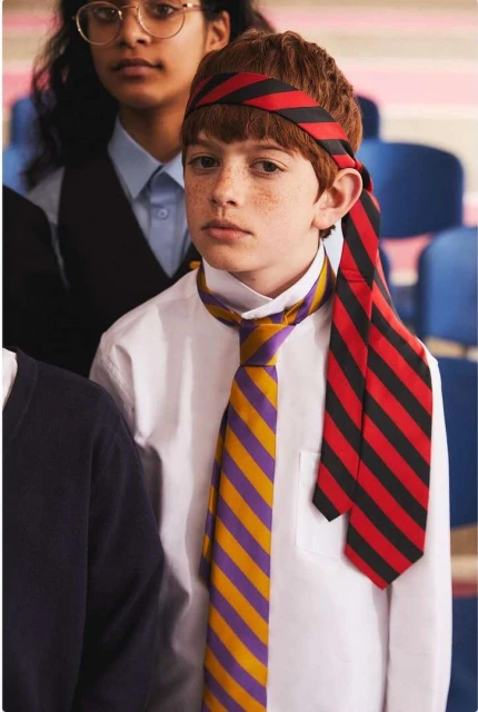 Young boy with red hair and freckles wearing a striped head band and a tie around his neck, standing in a classroom setting.