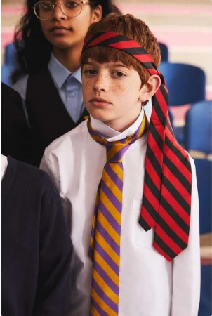 Young boy with red hair and freckles wearing a striped head band and a tie around his neck, standing in a classroom setting.
