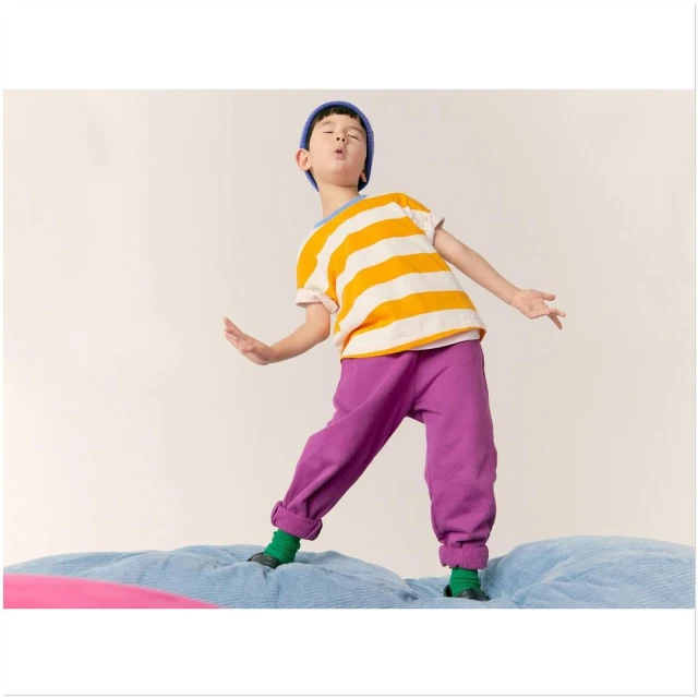 Young boy with a blue knit cap and a yellow striped shirt stands on a blue mattress in a studio setting, striking a fun, playful pose.