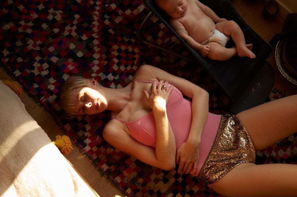 Woman in pink top and gold shorts lays on colorful rug indoors with a baby in black seat in the background in natural light.