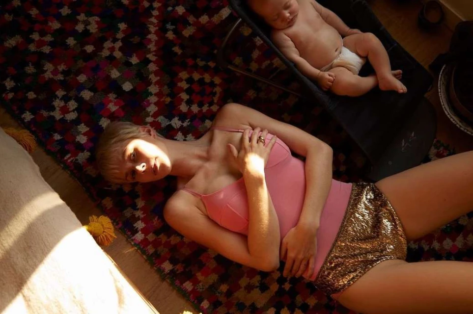 Woman in pink top and gold shorts lays on colorful rug indoors with a baby in black seat in the background in natural light.