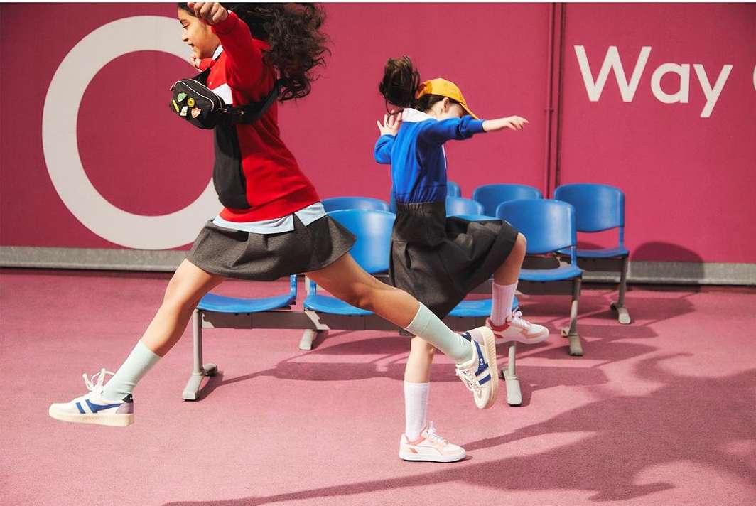 Two children leap in a playful manner inside, wearing school uniforms and sneakers. The background features a pink wall and blue chairs.