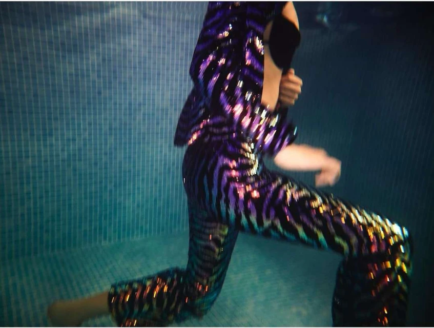 Adult wears a vibrant, sequined pantsuit underwater in a pool; the attire shimmers against a backdrop of blue tiled walls.