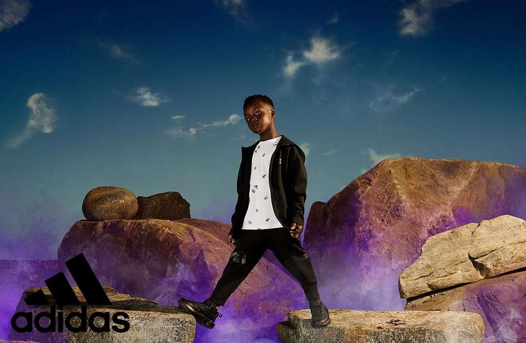 A Black child poses on rocks in Adidas brand apparel against a vibrant sky backdrop, showcasing activewear in an outdoor setting.
