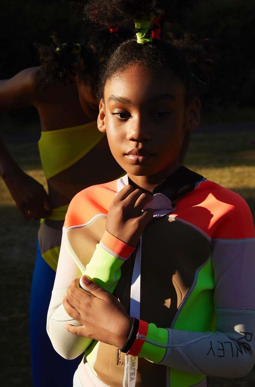 Two young African-American girls wearing athletic apparel posing outdoors. One girl faces forward, while the other is partially obscured.