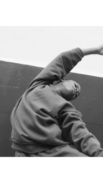 A child is stretching and bending backward outdoors in a black-and-white studio shot, wearing a sweatshirt against a neutral wall.