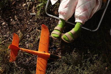 Child seated in folding chair wearing quilted trousers and green socks, with orange toy airplane on grass