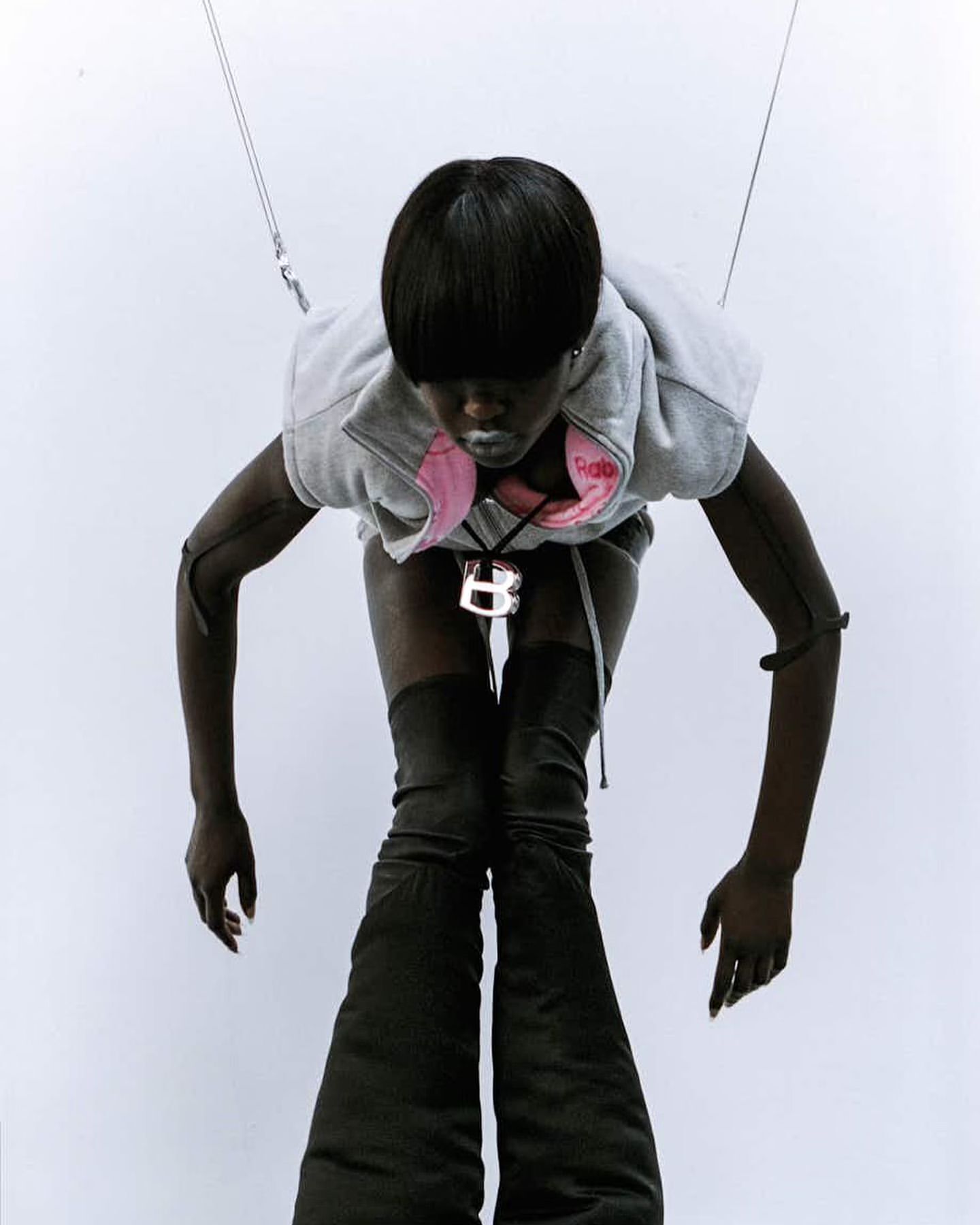 Woman with short black hair wears a grey hoodie vest and high, black, wide-legged boots against a white background in a fashion shot.